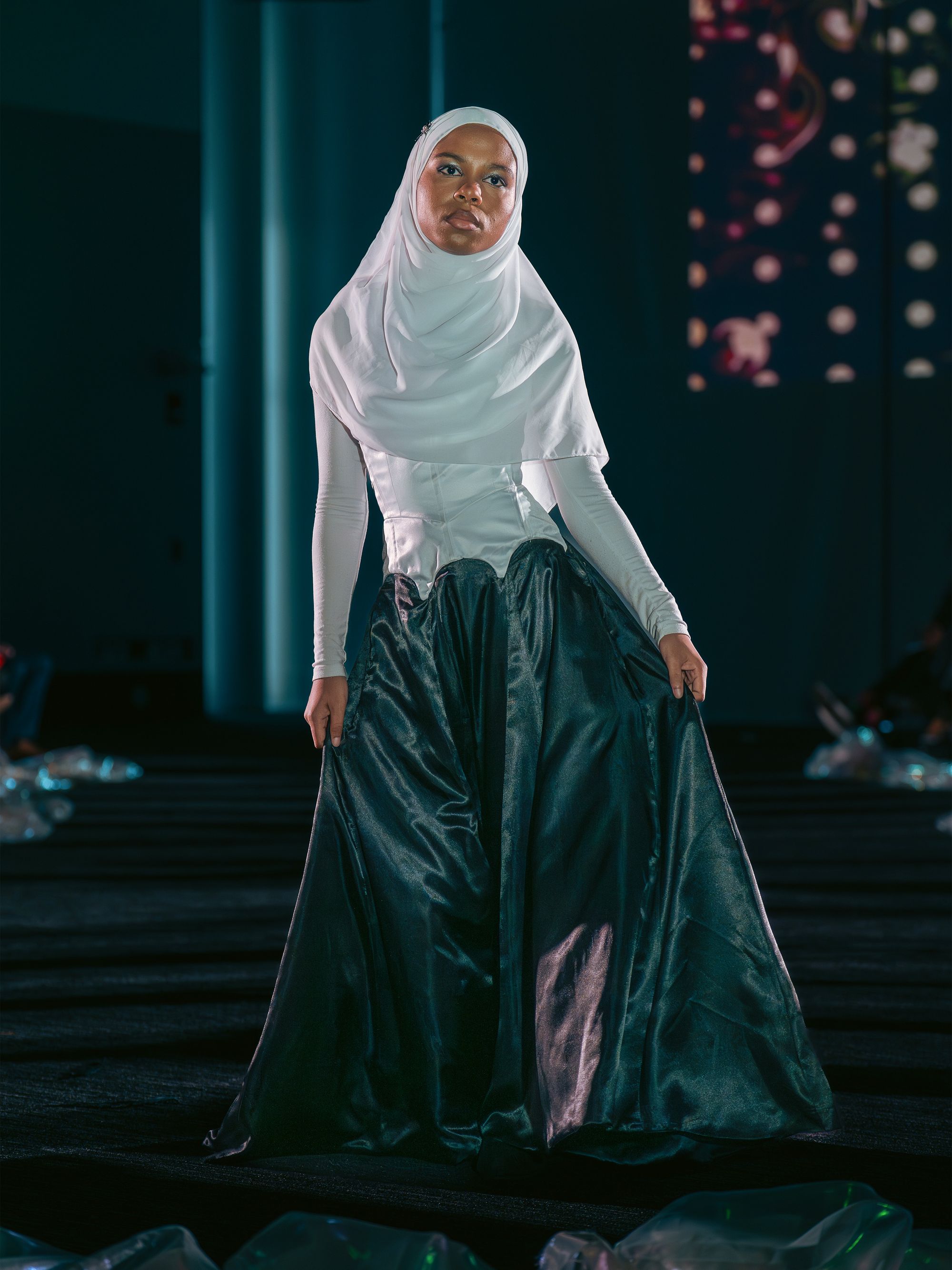 Model in a white hijab and long-sleeved top walks confidently in a high-waisted, dark teal satin skirt with a sculpted white corset, holding the skirt with both hands.